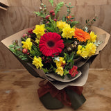 A bouquet of multicolored flowers including yellow, red, and orange flowers, with green foliage and red ribbon, wrapped in brown paper.