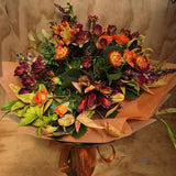 A floral bouquet featuring a variety of flowers in shades of orange, red, green, and yellow, wrapped in brown paper with ribbon.