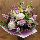 A floral bouquet featuring a variety of flowers in pastel tones, including white, green, and pink, wrapped with mauve and white paper.