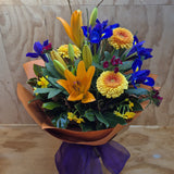 Bouquet of flowers with orange, yellow, and blue flowers wrapped in orange paper with a purple ribbon.