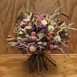 A dried flower bouquet with purple, pink, cream, and green botanicals arranged in a rounded shape, set against a light wood background.