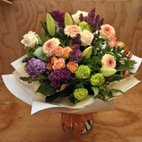 A bouquet of flowers featuring a mix of pastel tones including peach, green, purple, and white colors, wrapped in paper.
