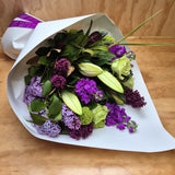 A bouquet of flowers featuring a mix of purple, white, and green colours, wrapped in white paper.