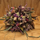 A dried flower bouquet with purple, pink, green, and brown botanicals arranged on a wooden surface against a wood-paneled background.