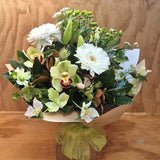 A mixed floral bouquet featuring white gerbera daisies, light green orchids, white helebores, Chrysanthemums and green foliage, wrapped in brown paper with a sheer olive-green ribbon, set against a wooden background.