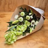 A fresh flower bouquet wrapped in brown kraft paper, featuring light green cymbidium orchids, white lilies, freesias, gerberas and green foliage, displayed on a wooden surface.