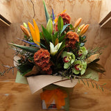 A vibrant mixed floral arrangement featuring orange chrysanthemum and lilies, tropical birds of paradise green orchids, and lush green foliage, wrapped in brown paper in a water-box with an orange ribbon, set against a wooden background.