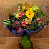 A floral bouquet featuring a variety of flowers including shades of yellow, red, purple and green, arranged in a wrap with purple and brown tones.