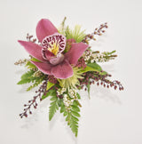 A floral wristlet or boutonniere featuring a prominent pinkish-purple orchid with a marbled yellow and burgundy center, surrounded by green ferns, small white flowers, and burgundy berry sprigs on a light white background.