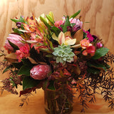 A colourful floral arrangement in a glass vase with a variety of flowers including freesias and green succulents, set against a wooden background.