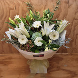A floral bouquet of white lilies, gerbera, green succulents, and alstroemaria wrapped in white paper with a soft green ribbon, displayed on a wooden surface against a wood-paneled background.