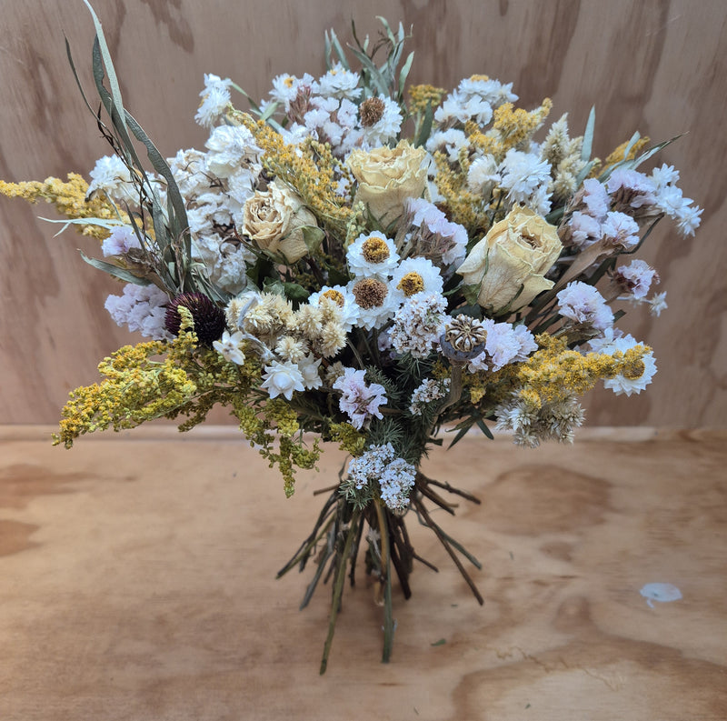 A dried floral bouquet featuring a mix of soft creamy shades, with white and a touch of yellow flowers, arranged in a naturalistic style on a wooden surface.