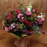 A bouquet of flowers with a variety of colors including white, purple, burgundy and green, wrapped in brown paper with a pink ribbon.