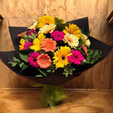 bouquet of yellow, hot pink and apricot gerberas with foliage in a bouquet, wrapped in black paper.